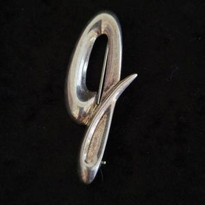 May include: A polished silver-tone metal brooch shaped like a stylized, elongated letter Q. The design features smooth, curved surfaces with a reflective finish and a pointed tail extending to the right, with a pin mechanism visible on the back.