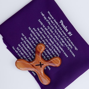 Op de afbeelding: Een houten kruis met een kruisvormige uitsparing rust op een paarse doek. De doek bevat de tekst van Psalm 91 in het wit. Het kruis is lichtbruin met een glanzende afwerking.