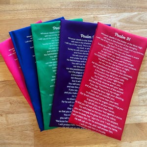 Op de afbeelding: Een stapel kleurrijke stoffen bladwijzers, elk met een andere kleur en een deel van Psalm 91. De bladwijzers zijn in roze, blauw, groen, paars en rood. De tekst is in het wit gedrukt.