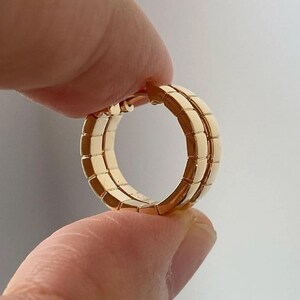 Pu&ograve; includere: Un orecchino a cerchio color oro con un design rettangolare. L'orecchino &egrave; tenuto tra due dita su uno sfondo bianco. L'orecchino ha un design a doppio strato.