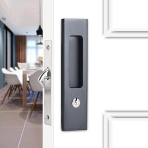 May include: A dark gray recessed door handle with a keyhole and a long, rectangular grip. A silver-colored door latch is visible on the left side. The handle is mounted on a white door, with a blurred interior in the background.