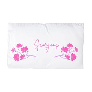 May include: White pillowcase featuring the word "Georgong" in pink script. Pink floral branches with purple stems are on either side of the text. The pillowcase is rectangular.
