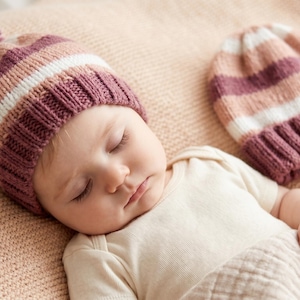 Puede incluir: Un bebé dormido con un gorro de punto a rayas con un pompón. El gorro presenta tonos de rosa, blanco y morado. Un segundo gorro a juego está al lado del bebé. El bebé lleva un body blanco.