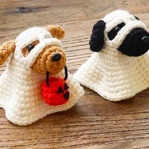 May include: Two crocheted dog figurines dressed as ghosts. One dog is light brown with a small red pumpkin bucket in its mouth. The other dog is black and white. Both are wearing white crocheted ghost costumes that cover their bodies and heads.
