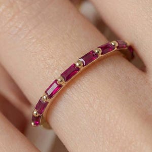 May include: A gold band ring featuring a series of baguette-cut red rubies set in a row. Each ruby is separated by a small gold bead, creating a continuous line of gemstones along the top half of the thin, polished yellow gold metal band.