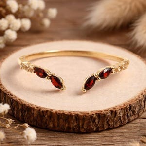 May include: A gold open-style ring featuring two marquise-cut deep red garnet gemstones on each end, accented by small clear stones along the band. The ring has a polished metallic finish and a split design that leaves an opening at the front.