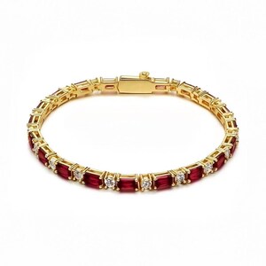 Puede incluir: Una pulsera de tenis dorada con rubíes rojos de corte baguette y diamantes blancos redondos alternados en una banda metálica continua. La pieza incluye un mecanismo de cierre rectangular seguro en la parte superior del aro.