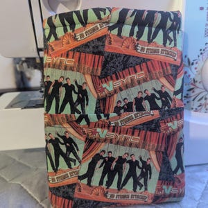 Op de afbeelding: Een rechthoekige stoffen hoes met een herhalend patroon van een boyband op een podium met de tekst "NSYNC" en "NO STRINGS ATTACHED". De hoes heeft een zwart, rood en groen kleurenschema.