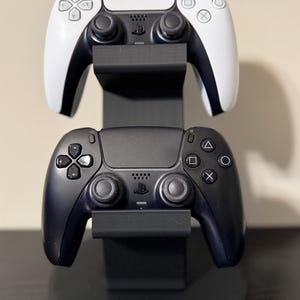 May include: A black and white gaming controller sits atop a black controller. The controllers are on a black stand. The top controller is white with black accents. The bottom controller is black.