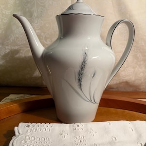 May include: White porcelain coffee pot with a lid and handle, featuring a wheat stalk design in blue and gray. The pot has a silver trim and a curved spout. A white eyelet lace napkin is in the foreground.