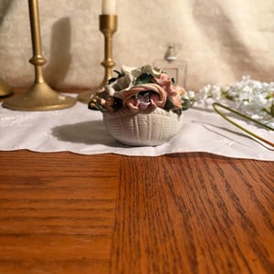 May include: A small, white ceramic basket filled with colorful, sculpted flowers. The basket has a woven texture and is filled with pink, white, and green flowers. Two gold candle holders and a white candle are in the background.