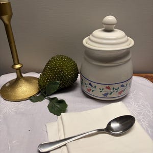 May include: A white ceramic jar with a lid, decorated with blue and red floral patterns, sits on a white embroidered cloth. A silver spoon and folded white napkins are in the foreground. A gold candlestick and a green decorative object are also present.