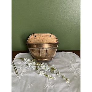 May include: A semi-circular wall-mounted planter made of hammered brass with a polished finish. The piece features a decorative rope-like trim detail across the front and two keyhole slots at the top for mounting. A sprig of white flowers lies below.