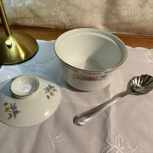 May include: A white porcelain sugar bowl with a floral design and gold trim, accompanied by a matching lid and a silver spoon. The lid and bowl feature a delicate pattern of blue, pink, and yellow flowers.