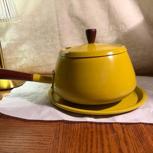 May include: A vintage yellow enamel saucepan with a matching lid and saucer. The saucepan has a wooden handle and a dark brown wooden knob on the lid. The set is placed on a white doily.