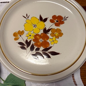 Puede incluir: Plato de cerámica color crema con diseño floral. Las flores son amarillas, naranjas y marrones, con hojas marrones oscuras. El plato tiene un borde dorado y un borde marrón.