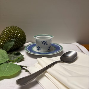 May include: A small, white teacup with blue leaf designs sits on a matching saucer with blue and green trim. A stainless steel spoon rests on a stack of folded white napkins, with a green decorative fruit and leaves nearby.
