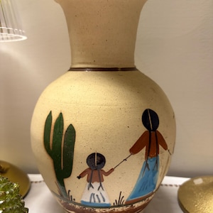May include: Tan ceramic vase with a wide base and flared top. The vase features a hand-painted design of a cactus and figures in traditional clothing. The vase is approximately 10 inches tall, perfect for displaying flowers or as a decorative piece.