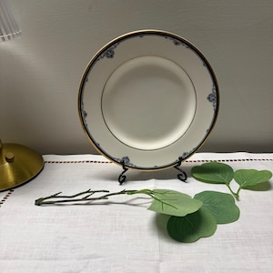 May include: A white plate with a gold rim and blue floral accents, displayed on a black metal stand. The plate is set on a white tablecloth, with green leaves and a branch adding a touch of nature.