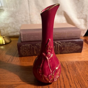 May include: A burgundy-colored ceramic vase with a gold rim and gold leaf design. The vase is tall and slender, with a flared opening. It is sitting on a wooden surface.