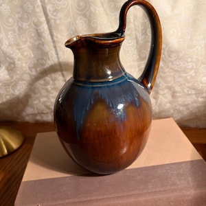 May include: A ceramic pitcher with a dark brown and blue glaze. The pitcher has a rounded body, a curved handle, and a spout. It is sitting on top of a stack of books. The pitcher is a decorative item.