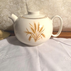 May include: A white ceramic teapot with a lid and handle. The teapot features a gold band around the rim and a wheat stalk design in shades of gold and brown. The teapot is sitting on a white cloth.