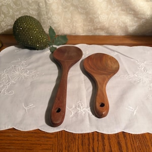 May include: Two wooden spoons with a rich brown hue, each featuring a rounded bowl and a long handle with a hole for hanging. They rest on a white, embroidered cloth, suggesting a kitchen or dining setting.