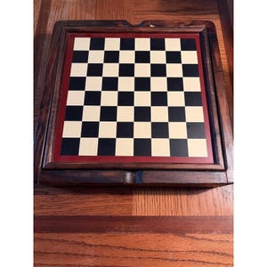 May include: A wooden chess board with a black and cream checkered playing surface. The board is framed in dark brown wood with a red border. The board is set on a wooden surface.