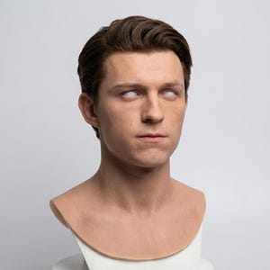Pu&ograve; includere: Un busto in silicone realistico di una giovane persona con capelli castani corti e acconciati, pelle chiara e occhi completamente bianchi. La scultura cattura dettagliati tratti del viso, inclusi naso, labbra e orecchie definiti su un supporto bianco.
