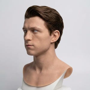 Pu&ograve; includere: Un busto in silicone realistico di una giovane persona con capelli corti castano scuro pettinati con riga laterale. La figura presenta una pelle chiara, un'espressione facciale neutra e occhi bianchi solidi e privi di tratti, su base bianca.