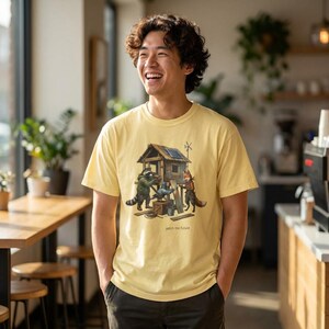 Può includere: T-shirt giallo chiaro con un'illustrazione a cartoni animati di una casa di legno con un pannello solare e una turbina eolica. Un procione e una volpe lavorano sulla casa. Il testo "patch the future" è sotto.