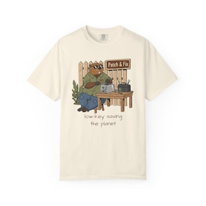 Pode incluir: Camiseta creme com um castor de desenho animado usando óculos de sol, consertando uma torradeira. O castor está em frente a uma cerca de madeira com uma placa "Patch & Fix". O texto "low-key saving the planet" está abaixo.
