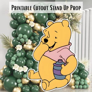 Puede incluir: Un accesorio recortado de Winnie the Pooh amarillo con una camisa rosa y un tarro de miel. El accesorio está frente a un arco de globos con globos verdes y dorados. El texto "PRINTABLE CUTOUT STAND UP PROP" está en la parte superior.