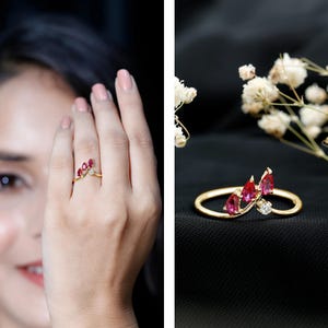 May include: A gold ring featuring three pear-shaped red gemstones arranged in a cluster, accented by a single small round clear gemstone on a thin, curved band. The design follows a V-shaped silhouette, showcasing a polished metallic finish.