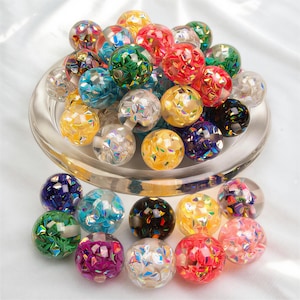 May include: A selection of multi-coloured, iridescent glass beads. The spherical beads are in shades of red, yellow, blue, and green, with a faceted, sparkling finish. They are displayed on a clear glass plate. The beads are approximately 2 cm in diameter.