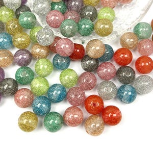 May include: A selection of colourful, glittery beads in shades of gold, green, pink, and blue. These round beads are scattered, highlighting their sparkling texture. Ideal for jewellery making or craft projects.