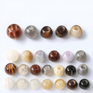 May include: A collection of twenty-three spherical beads arranged in four rows. The beads feature various finishes including translucent, opaque, marbled, and speckled textures in shades of brown, gray, cream, white, and black with central holes.