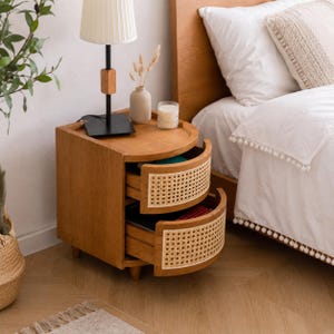 May include: A wooden nightstand with two curved drawers featuring light-colored woven cane panels. The top surface holds a black lamp with a white pleated shade, a small beige vase with dried stems, and a clear glass candle holder with wax.