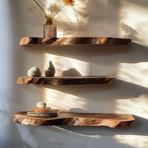 Pode incluir: Tr&ecirc;s prateleiras de parede horizontais feitas de madeira natural com bordas r&uacute;sticas. A prateleira superior cont&eacute;m um vaso de vidro com flores amarelas. A prateleira do meio cont&eacute;m duas pedras lisas. A inferior cont&eacute;m livros e uma pedra.