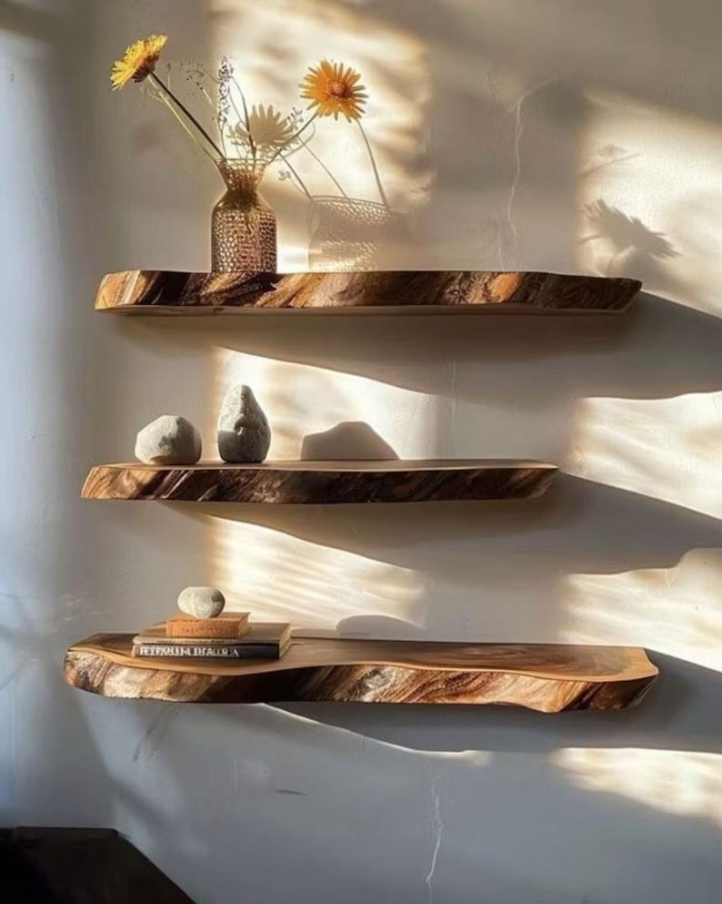 Pode incluir: Tr&ecirc;s prateleiras de parede horizontais feitas de madeira natural com bordas r&uacute;sticas. A prateleira superior cont&eacute;m um vaso de vidro com flores amarelas. A prateleira do meio cont&eacute;m duas pedras lisas. A inferior cont&eacute;m livros e uma pedra.
