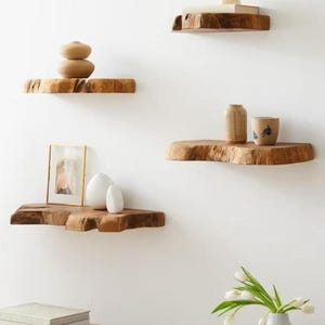 Pode incluir: Quatro prateleiras flutuantes montadas na parede feitas de madeira natural com bordas irregulares. As prateleiras est&atilde;o dispostas em um padr&atilde;o escalonado e cont&ecirc;m v&aacute;rios itens decorativos, incluindo uma moldura dourada, vasos brancos e uma x&iacute;cara.