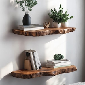 Pode incluir: Duas prateleiras horizontais montadas na parede feitas de madeira natural com bordas irregulares. A prateleira superior cont&eacute;m uma pilha de livros, um vaso preto com folhagem verde, uma pequena escultura de madeira e dois vasos brancos.