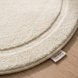 May include: A circular off-white rug with a thick, textured pile and a raised border detail. A small white fabric tag is attached to the edge of the rug, featuring the printed black text: 100% Wool. Hand Tufted. The rug has a soft, dense appearance.