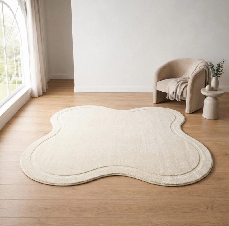May include: An off-white area rug with a distinct four-lobed, clover-like shape and a smooth, continuous border line following the perimeter. The rug features a soft, uniform texture and is laid flat on a light-colored wooden floor surface.