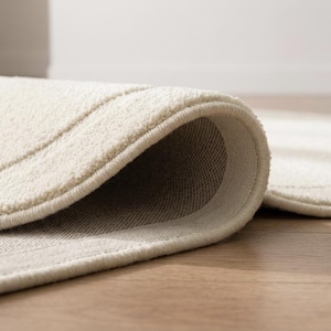 May include: A cream-colored area rug with a soft, textured pile surface and a folded corner revealing a woven backing. The rug features a subtle linear pattern embossed into the fabric, with finished edges and a consistent light neutral tone.
