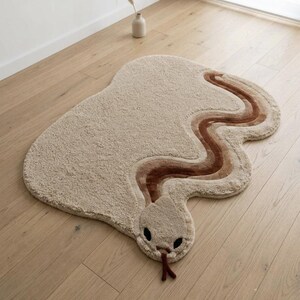 以下が含まれることがあります： ヘビの形をしたベージュ色のエリアラグで、表面はテクスチャー加工されています。ラグには茶色の色合いでうねるような体の模様が描かれており、丸みを帯びた頭部には暗い楕円形の目があり、前縁からは暗い茶色の二股に分かれた舌が突き出ています。