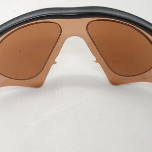 May include: Black sunglasses with brown tinted lenses. The sunglasses have a curved black frame with a silver accent on the temples. The lenses are a light brown colour and are shaped to fit the frame.