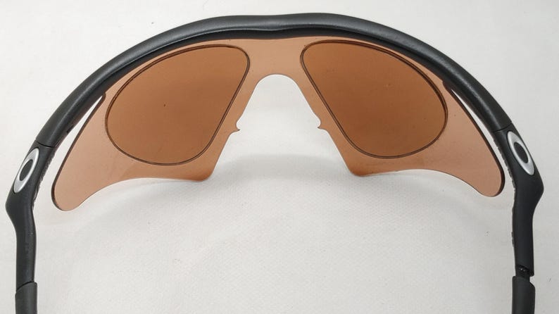May include: Black sunglasses with brown tinted lenses. The sunglasses have a curved black frame with a silver accent on the temples. The lenses are a light brown colour and are shaped to fit the frame.