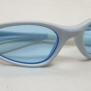 K&ouml;nnte beinhalten: Hellgraue Sonnenbrille mit blauen Gl&auml;sern. Die Fassungen haben ein geschwungenes Design mit einer einzigartigen Form an den B&uuml;geln. Die Gl&auml;ser sind hellblau, und der Gesamtstil ist modern und elegant.