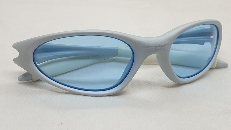 K&ouml;nnte beinhalten: Hellgraue Sonnenbrille mit blauen Gl&auml;sern. Die Fassungen haben ein geschwungenes Design mit einer einzigartigen Form an den B&uuml;geln. Die Gl&auml;ser sind hellblau, und der Gesamtstil ist modern und elegant.